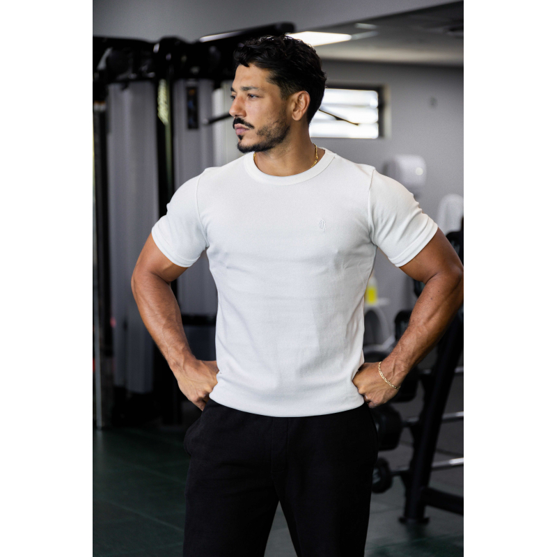 Homme musclé en t-shirt ESSENTIAL blanc, debout en salle de sport, poings sur les hanches, regard sur le côté.