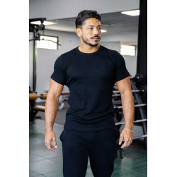 Homme musclé portant t-shirt noir ESSENTIAL noir, en salle de sport, regard vers la droite, cheveux foncés et barbe légère.