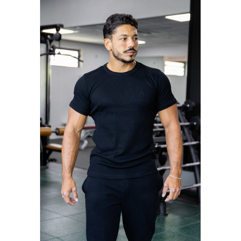Homme musclé portant t-shirt noir ESSENTIAL noir, en salle de sport, regard vers la droite, cheveux foncés et barbe légère.