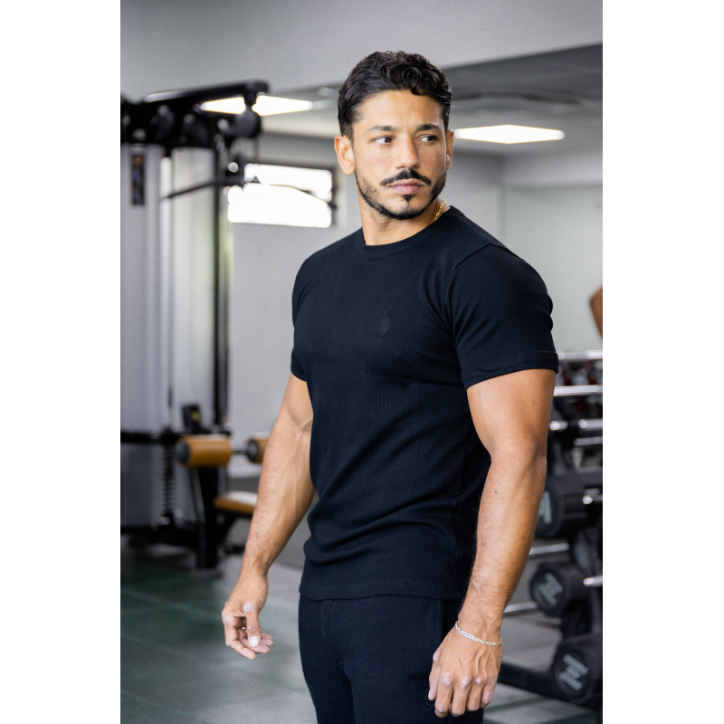 Homme musclé en t-shirt noir ESSENTIAL, debout dans une salle de sport, muscles visibles et expression concentrée.