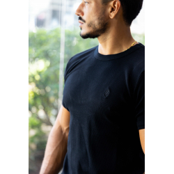 Homme portant t-shirt noir ESSENTIAL, vue de profil, près d’une fenêtre, cadenas doré au cou, petit logo embossé sur le torse.