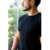 Homme portant t-shirt noir ESSENTIAL, vue de profil, près d’une fenêtre, cadenas doré au cou, petit logo embossé sur le torse.