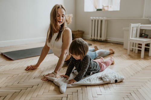 Une mère souriante et sa jeune fille font des exercices de yoga/étirements sur un parquet clair, dans une pièce lumineuse.