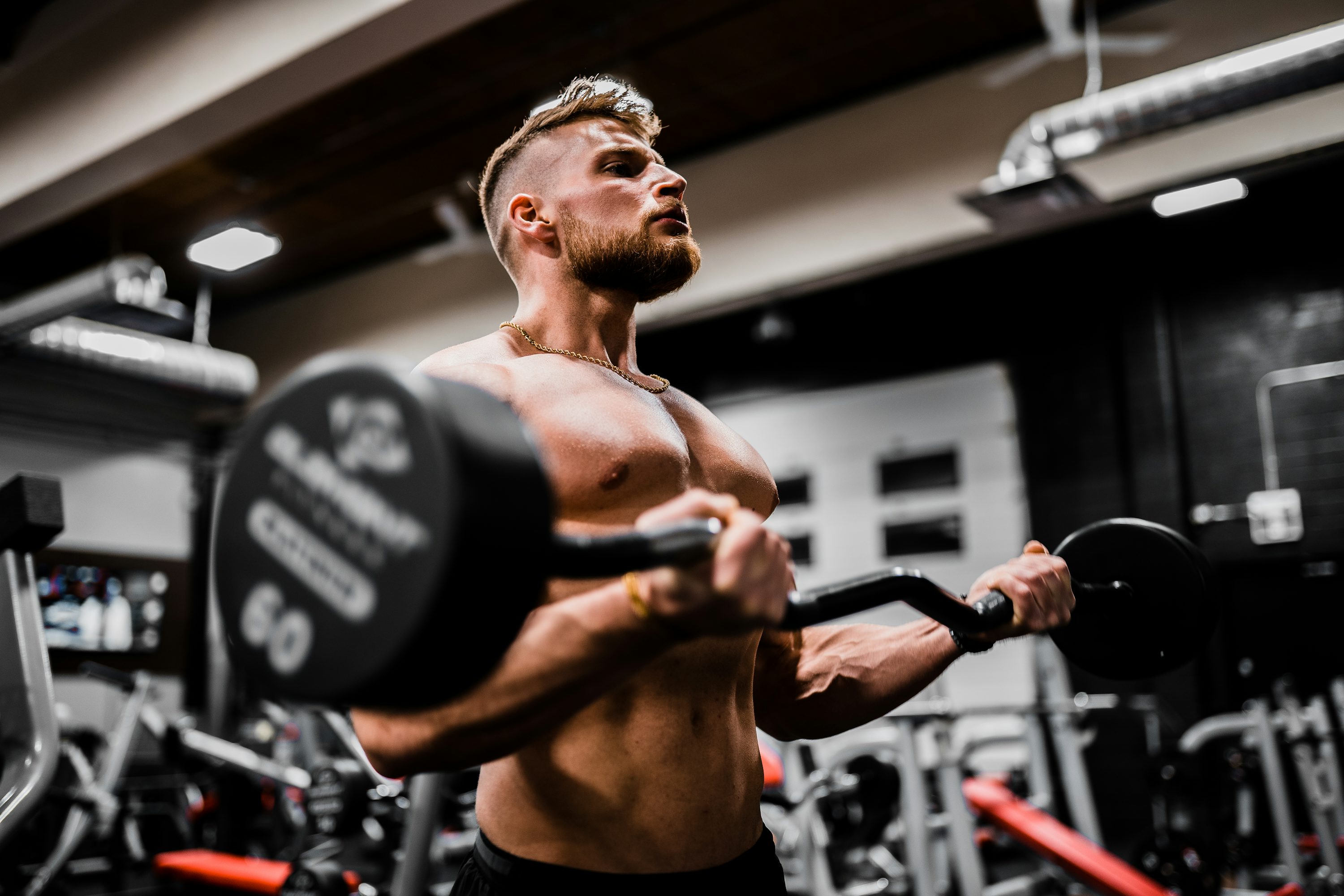 Homme musclé effectuant un curl avec barre guidée en salle de sport, demi-tampleur, muscles tendus, ambiance gym au fond.