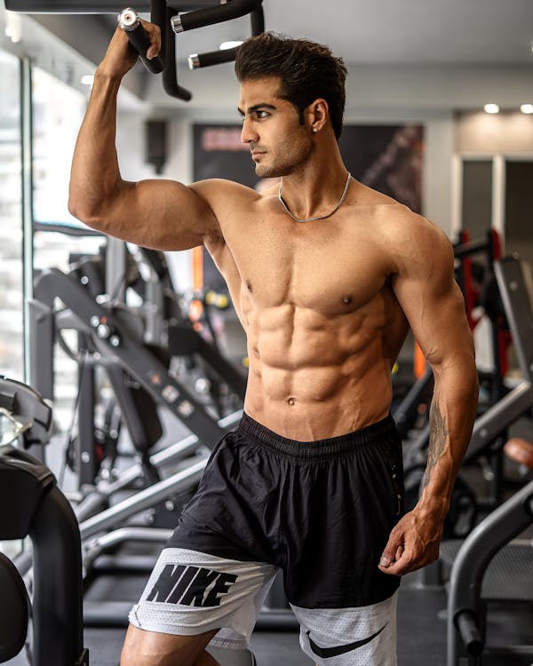 Homme musclé en train de faire un exercice d’épaule au gym, portant short noir et gris, en salle de musculation.